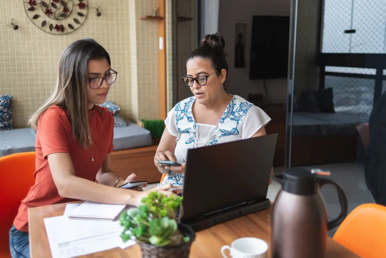 Mother and daughter doing finances together at home. They talk and handle some bills, looking and pointing at laptop screen