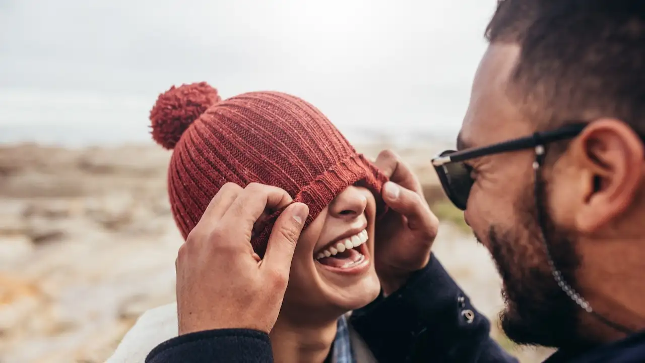 Loving couple having fun outdoors. Man covering eyes of woman with cap.