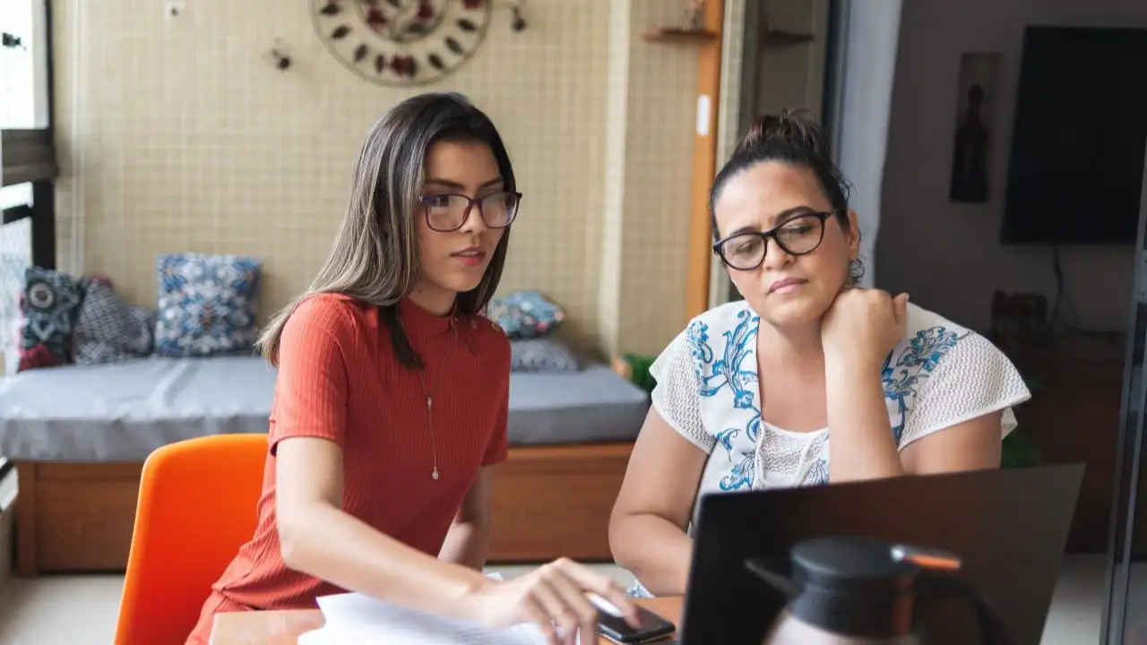 Mother and daughter doing finances together at home. They talk and handle some bills, looking and pointing at laptop screen