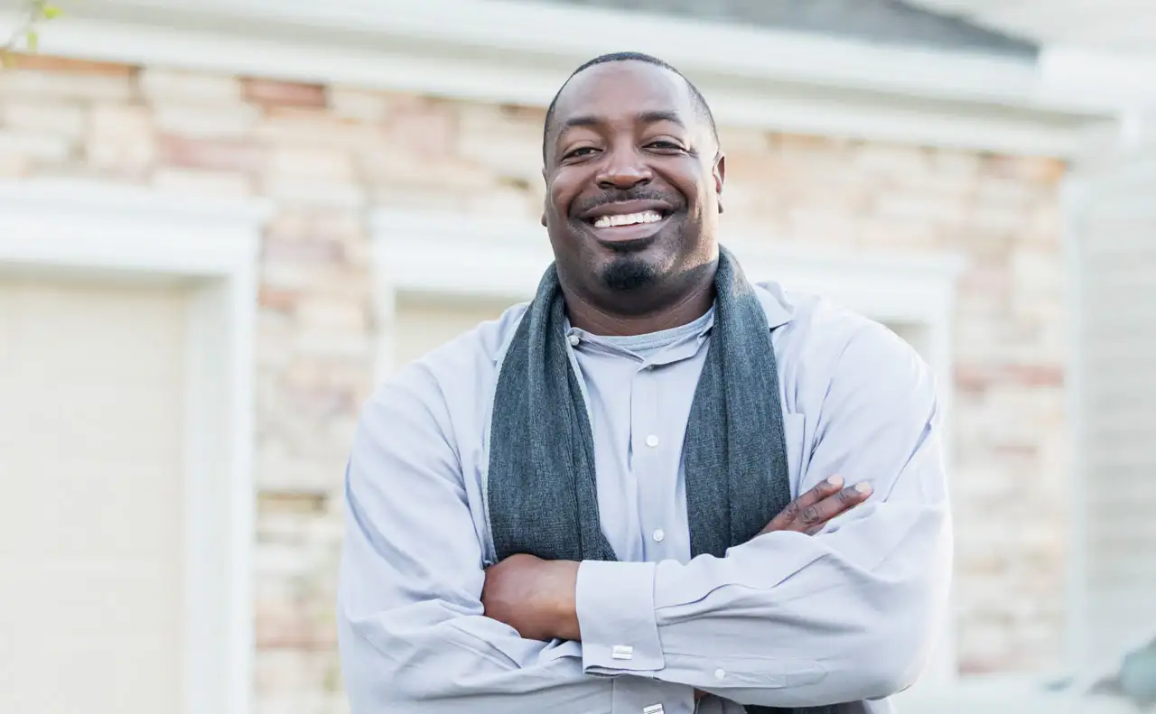 Young Afro-American man with his hands folded standing in front of his home. He has a big smile.