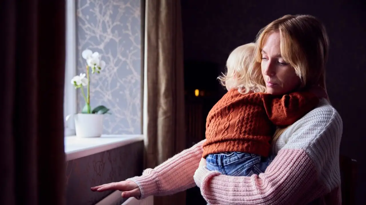 Mother With Son Trying To Keep Warm By Radiator At Home During Cost Of Living Energy Crisis