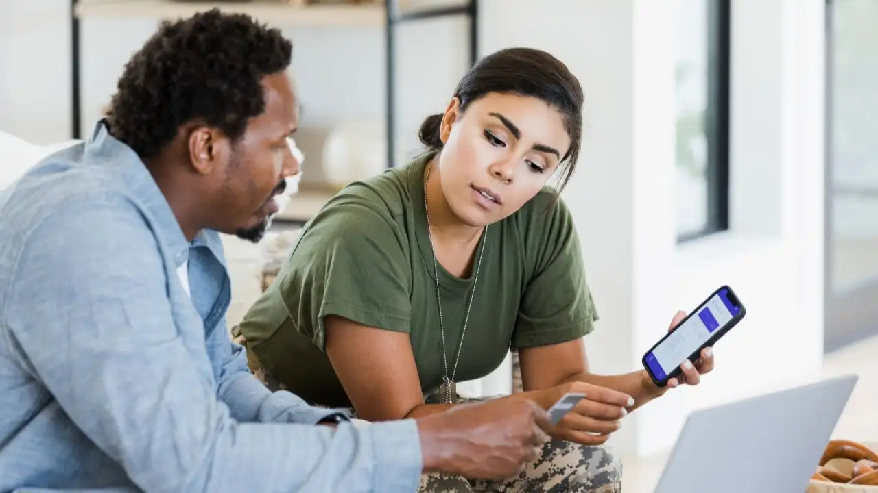 The mid adult man holds a credit card as he and his soldier wife discuss home finances.